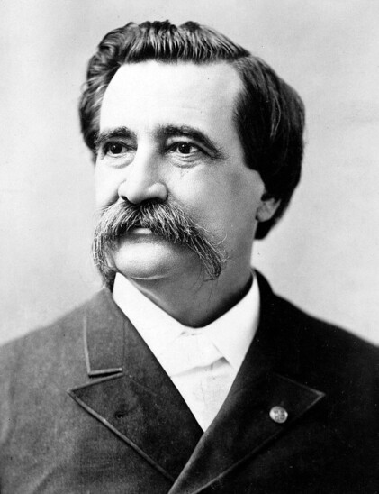 Black and white photo of John A. Logan. He is wearing a dark suit coat and collared shirt with no tie. He has a mustache that puts all other mustaches to shame; it extends below his jaw-line.