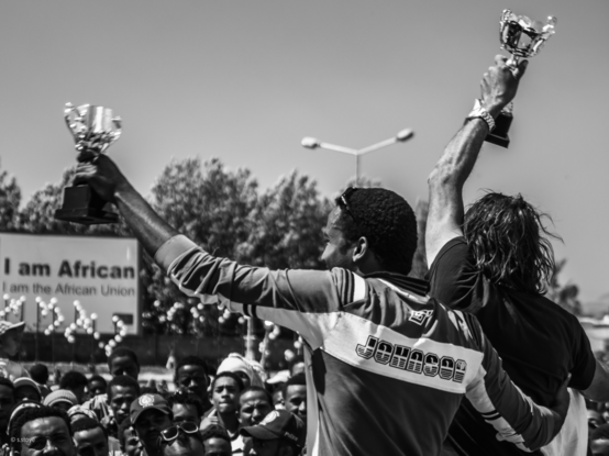 black and white photography of a street scene in addis ababa. Two men in sports jerseys each hold up their trophies with their left arms at an awards ceremony. in the background audience (only the heads are visible). on the back left is an advertising poster with the inscription “i am african”. the photo was taken after a car race in the city.
