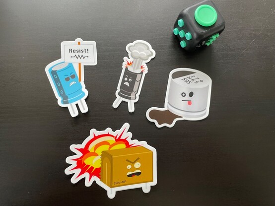 Four of Geekman's "Bad Caps" capacitor cartoons as stickers (fidget cube for scale)