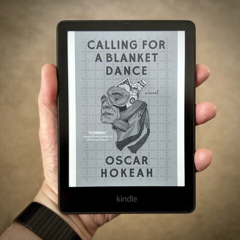 Hand holding a Kindle Paperwhite e-reader showing the cover of Oscar Hokeah's novel, Calling for a Blanket Dance