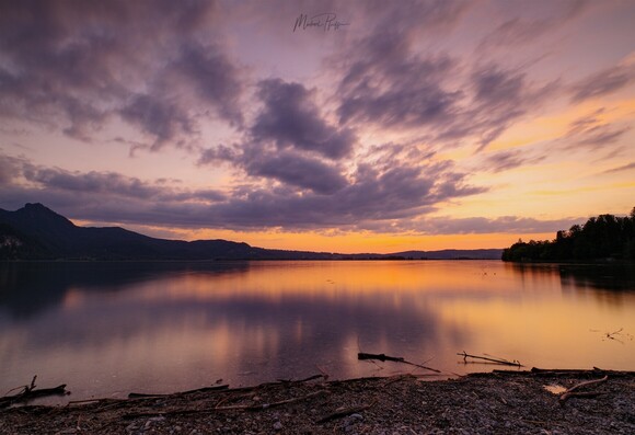 Im Vordergrund etwas Kiesstrand dahinter der See.Im Hintergrund das gegenüberliegende Ufer des Sees mit Bergen und Hügeln.Der Himmel orangerrot nach dem Sonnenuntergang mit einigen grauen Wolken im Wechsel. Das Bild ist langzeitbelichtet