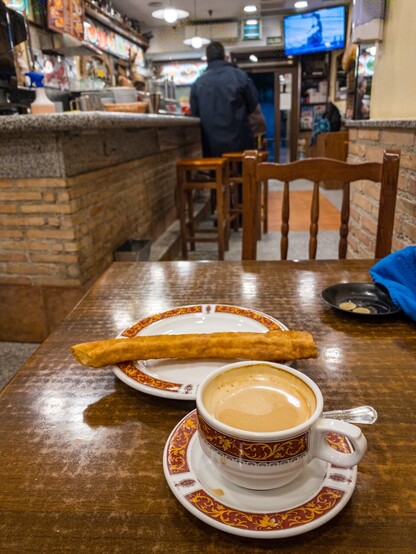 A cup of cafÃ© con leche and a cold, greasy churro on a table in a neighbourhood bar in Madrid, with some other customers watching the retirement of Real Madrid footballer Karim Benzema on the TV.