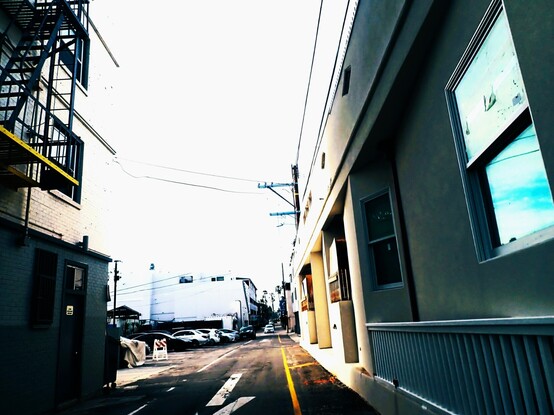 Los Angeles. A laneway. Between buildings. Blue sky reflection in window at right. The scene is somewhat bleak vibe, seedy, down on its luck, beautiful noirish perspective - (liminal space) In the distance white sky, white low rises, cars parked in a lot, wires, yellow line on road at right, parallex. Late afternoon. On left fire escape, white brick, large windows, greenish tones, a sense of taut space. March 2023.