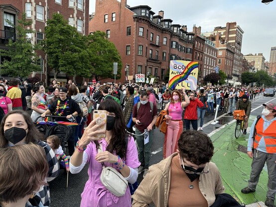 Marchers. In the foreground, one person takes a selfie. Someone else carries a rainbow covered sign reading "Bodily autonomy for all".