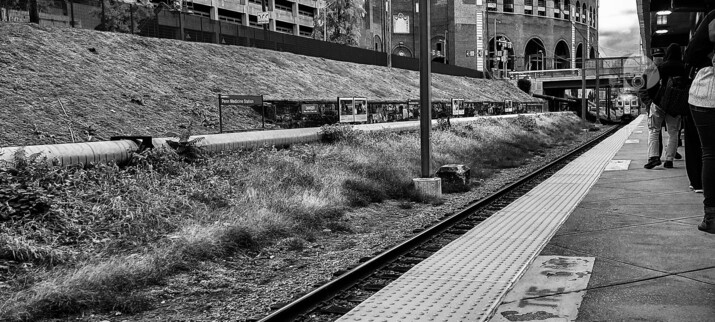 A black and white photo of a train approaching the station. A grassy embankment borders the tracks, atop it is a fence and a view of some brick buildings. A bridge can be seen in the background, just over the train. Passengers stand on the platform, waiting for the train to finish its approach for boarding.