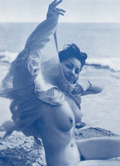 An old monochrome photograph of a woman lifting her gauzy dress to reveal her nude body. The cloth blows in the wind, and waves break in the background.
