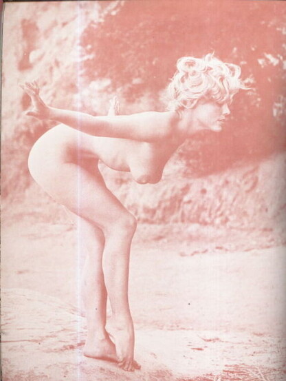 An old monochrome photograph of a nude woman striking an unusual ballet-like pose on a rocky beach. She is bent forward at the waist with arms extended out to the sides. Her right knee is bent and she is touching the ground with the tips of her toes.