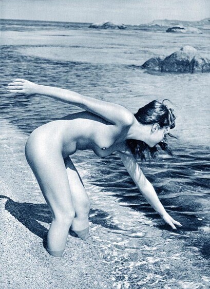 An old monochrome photograph of a nude young woman reaching down to dip her hand in the clear sea.