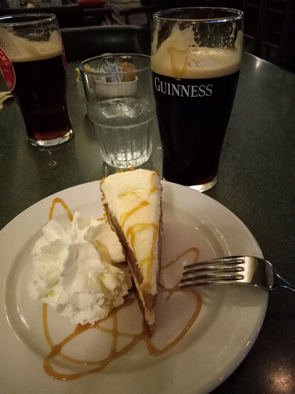 Set on a wooden table at a pub restaurant: a plate with a tall slice of carrot cake & fat dollop of whipped cream & two pints of brew (Guinness in the one, a Guinness & Smithwick's in the other).
