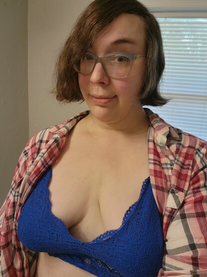 Standing in a room looking at you with trouble in their eyes is a nonbinary cutie with short brown hair, blue glasses, a sly smile, and a pinkish red flannel shirt. It is very open and upon further look is a blue lacy bra showing off very large boobs