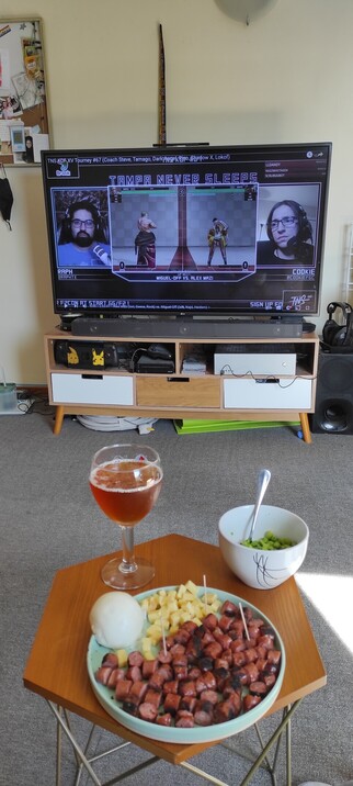 Interior photo. In the foreground a small table has a big glass of beer, a small bowl of soy beans (edamame) and a big plate with chopped cheese and sausages, as well as one big bean paste bun.

In the background, a big TV with a paused YouTube video. The video is  a Tampa Never Sleeps King of Fighters 15 weekly tournament VOD