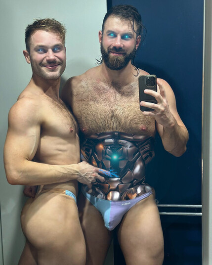 Two lean muscled, hairy droids embrace. One holds a camera and has long black hair, a beard, a furry chest, and a wrestler's build. The other is more lean with curly blond-brown hair, lighter chest hair, and is zapping electricity into the bigger bot. The bigger bot has its abdominal robotics exposed with a reactor starting to glow after the electricity is jolted into it.