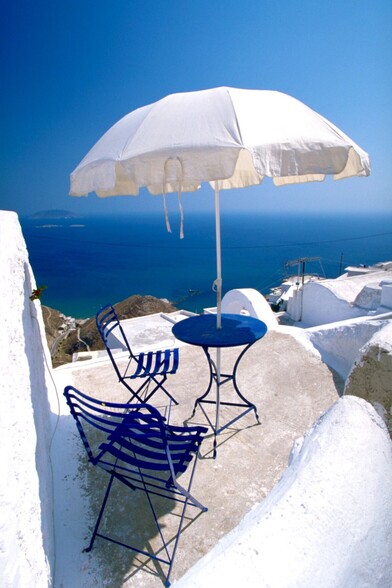 Vertical image of a small terrace in a sunny summer day overlooking the Mediterranean Sea. Walls are painted in white, same color as the sunshade. Metal chairs, two, and little round table are painted in dark Mediterranean blue.