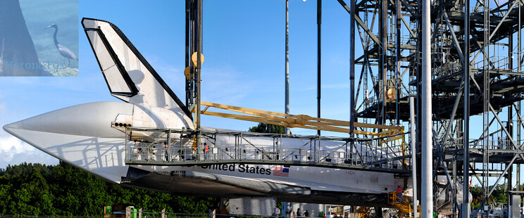 An outdoor, daylight photograph of NASAâ€™s Space Shuttle, a plane-like spacecraft, inside a rectangular truss-work structure with a crane attached to the top of the Shuttle Orbiter beginning to raise it. The Orbiter has a fuselage, wings, and vertical stabilizer of an aircraft and rocket engines for ascent and maneuver on-orbit, covered by a cone on the Shuttleâ€™s tail.