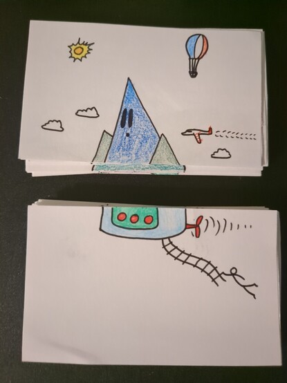 Two stacks of notecards. The top two cards combine to form a sketch of a mountain range riding through a cloudy sky on a blimp like craft, trailing a stick figure from a rope ladder.