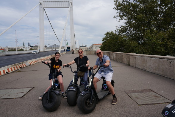 Three people sit astride big-wheeled scooters in Budapest. A road bridge is in the background.