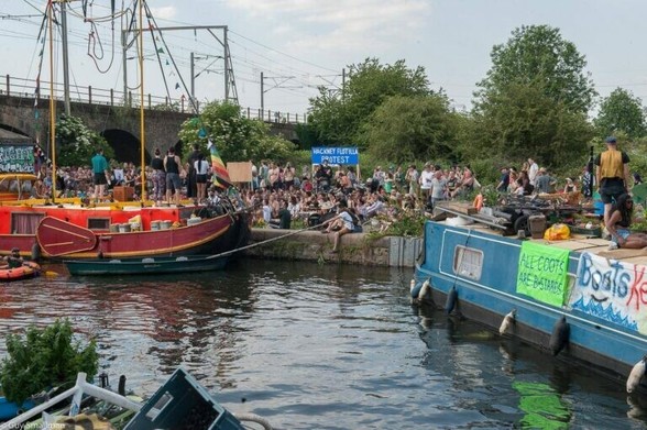 Narrowboats are moored by the bank, next to a railway line. There are banners on the boats reading ALL COOTS ARE BASTARDS. There are lots of people on the bank, and a banner reading HACKNEY FLOTILLA PROTEST