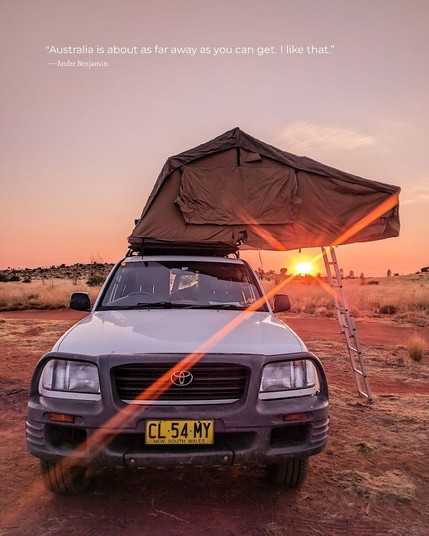 A closeup of the front profile of a small white truck, with a portable tent set up in the truck bed, with a ladder reaching down to the ground. The sun sets in the distance. The quote on the photo reads, “Australia is about as far away as you can get. I like that.” by Andre Benjamin.