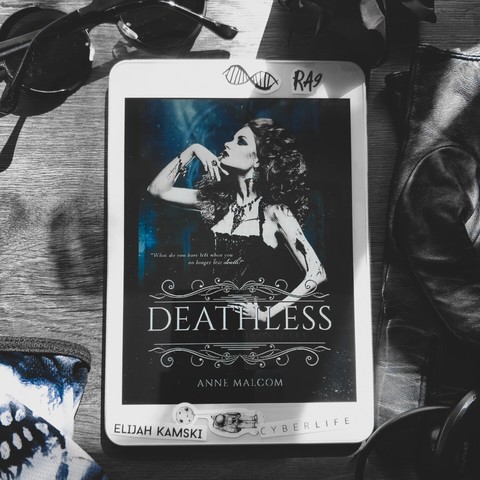 Deathless by Anne Malcom, second book in The Vein Chronicles series, ebook cover. It depicts a woman in a bodis type of a dress, long curly red hair, in some kind of church or gothic castle. White framed tablet is placed on a dark, greyish wooden table, with various dark items strewn around, such as round black sunglasses, leather gloves, a balled up piece of black paper, earphones, skull-print facemask.