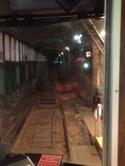 A view down a subway tunnel. Pillars on the left, colored green on bottom half, off white on top, converge in this view with the lights along the top right of the tunnel and the tracks below.  The window frame is visible at right, and at the bottom, a bit of the train's interior apparatus and a red placard.