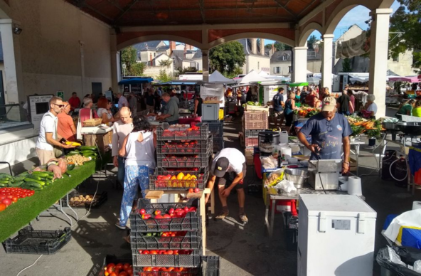 Part of a busy French market is undercover, with stalls arranged in lines under a roof supported by arches along two of its sides. The picture shows stalls and box crates laden with produce. The stall to the left displays tomatoes, cucumbers, peppers, sweet potatoes, potatoes and courgettes. Black box crates in the middle are stacked behind the stalls and contain red and yellow tomatoes. A stallholder to the right is wearing a dark blue t-shirt with a slogan from La Réunion. The island's food has French, Chinese, Madagascan, Mauritian, Indian & East African influences. He's surrounded by kitchen kit, is using a fryer and selling spice-scented curry.