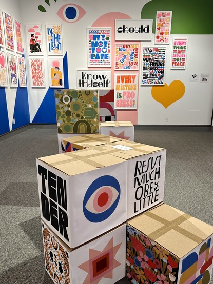 one of the rooms in Hold it Lightly, an exhibition of Lisa Congdon’s work at the St. Mary’s College Museum of Art. prints on the wall and on card board cubes in the middle of the room are illustrated in bright primary colors, and read “know thyself,” “TENDER,” “resist much, obey little” among others.