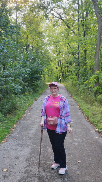 It's me. A woman with short pink hair, a pink cane, pale pink shoes, dark jeans, pink blue white flannel, pink trucker hat, pink hip pack, magenta T-shirt that says Outdoors Together in rainbow font. I'm standing on a trail in a park surrounded by greenery.