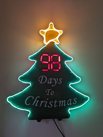 A christmas tree shaped wall hanging outlined in green led rope lighting with a yellow five pointed star on top. 

In the middle are two digital style numbers backlit in red above the white painted words 'Days to Christmas'

There are 98 days to go