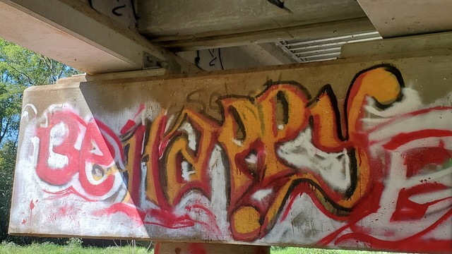 Attempted old english font graffiti, sprayed under a bridge on the horizontal supports. Undercoat sprayed in white. "Be" is in red. "Happy" is in yellow with black and red edges for contrast.
