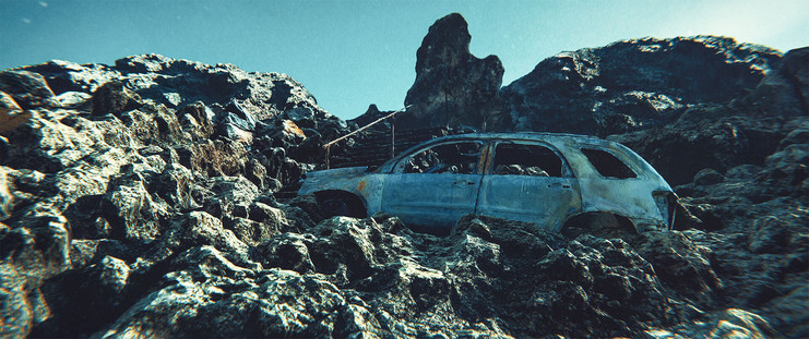 A car wreck halfway up a dry rocky slope. Beyond the car are some stairs to the summit of the hill.