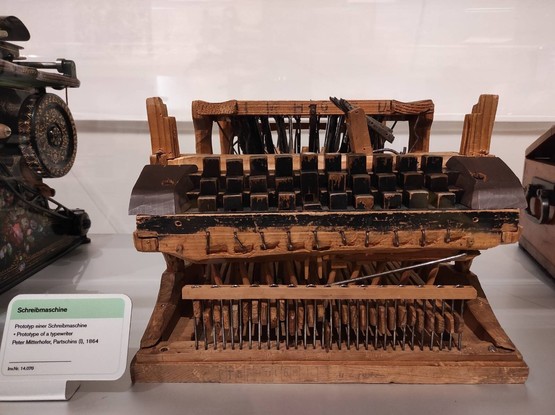 "Prototyp einer Schreibmaschine von Peter Mitterhofer aus Partschins" steht auf einer Tafel neben der Schrebmaschine. Die Schreibmaschine besteht aus Holz und Draht. Das Holz der dreißig Tasten ist schwarz gefärbt und blättert teilweise ab. Das Foto wurde im technischen Museum in Wien gemacht.