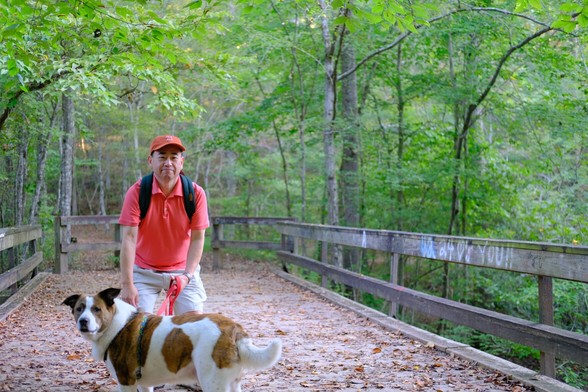 A man in an orange shirt and a cap and his white and brown dog on a wooden bridge with trees behind