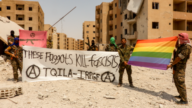 TQILA fighters in Raqqa with a banner that reads: These faggots kill fascists (with circle A’s), as well as a rainbow flag, and an anarchist banner with a machine gun and a circle A. By IRPGF, CC BY-SA 4.0, https://commons.wikimedia.org/w/index.php?curid=89781661