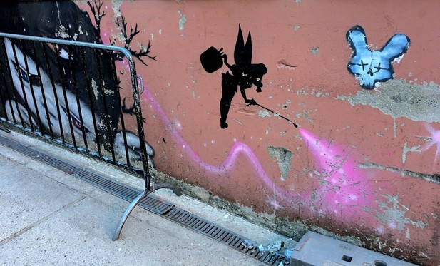 Graffiti of an elf spraying pink pixie dust on an orange wall. A metal fence.
