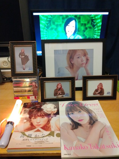 Kin-chan merch on a desk: Framed pictures, a pile of CDs, her two photobooks, and an AZALEA penlight. On a monitor on the background is the music video from her first song.