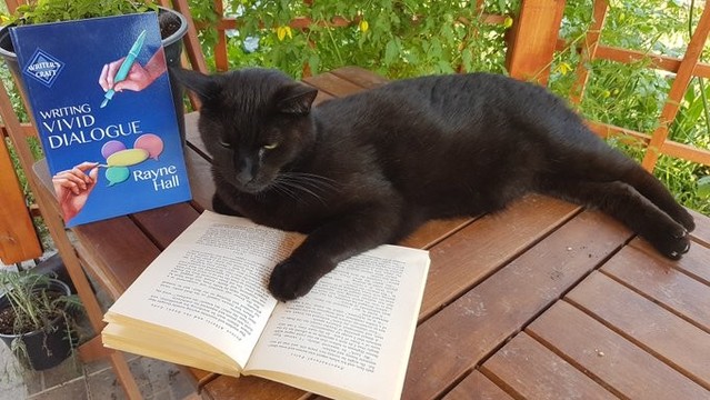 A cute  black cat lies on a wooden table. In front of him is an open book, and he has a paw on the pages, apparing to read the book. In the background is the book 'Writing Vivid Dialogue' by Rayne Hall.