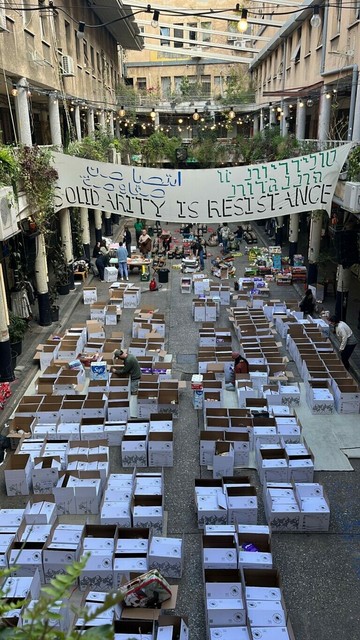 A photograph of a street in Tel Aviv with a banner over it saying SOLIDARITY IS RESISTANCE. There are boxes in the street that people are opening.