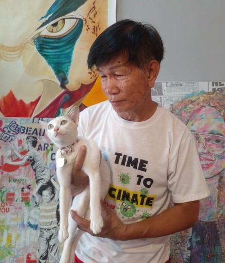 an asian man holding a white cat in an art studio, there are paintings behind them, he is wearing a white t-shirt