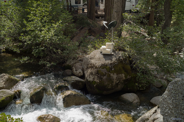 A TV Sattelite dish weighed down by light covered bricks on a rock by a stream with rushing water.   Portions of two travel trailers can be seen behind trees in the background.