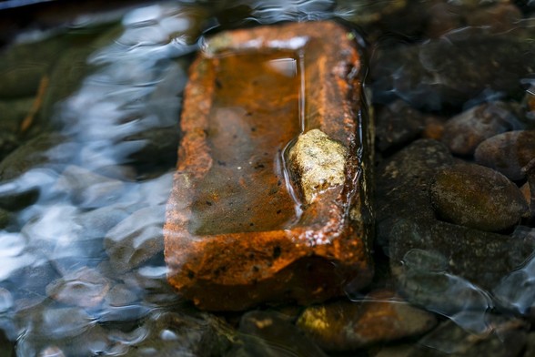 A single red brick sitting abandoned in a shallow creek. Ninety percent submerged in water with sunlight reflected off the rippling water.