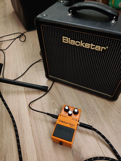Une pédale de distorsion Boss DS-1 de couleur orange, posée par terre devant un ampli Blackstar auquel elle est branchée.
Le sol est en parquet stratifié gris.