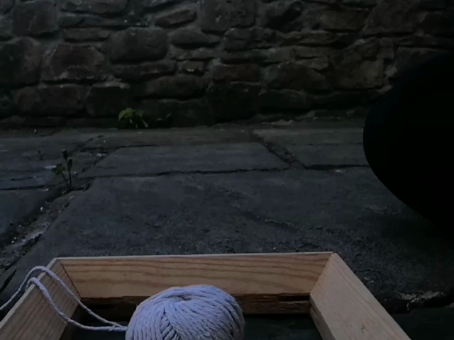A small square wooden picture frame and a ball of string on paving stones in an outdoor area. A white person who is mostly out of frame picks up both objects and wraps the string around the top edge of the frame, then places them back down again