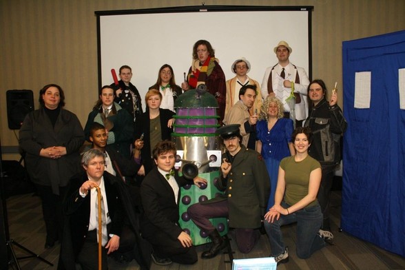 A cast of actors pose after a performance of a "Doctor Who" parody play in a hotel conference center panel room during a science-fiction convention portraying various incarnations of the Doctor, companions, rogues, and a Dalek and the TARDIS props.