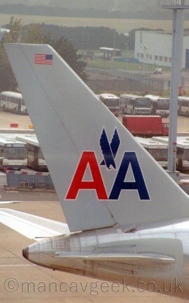 Closeup of the tail of a bare metal jet airliner with a pair of capital letter "A's" with a blue stooping eagle between them. taxiing from left to right at an airport, with rows of parked buses behind it.