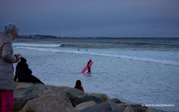 Photo of a surfer walking out the surf in the background with various passersby watching the waves and surfers. It is a cloudy and cold day.