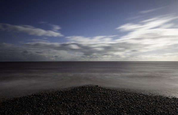 Standing on a pebbly beach staring out to sea. The beach is at your feet in a triangular pattern pointing out to sea. The waves are smoothed and the clouds are blurred from a long exposure.