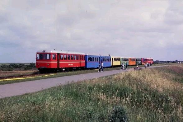 8 kunterbunte Triebwagenzüge und Beiwagen neben einer Landstraße auf eingleisiger Strecke und einem als Bahnsteig umfunktionierten Grünstreifen mitten auf dem mit Stoppelfelder umgebenden Platten Land. Ein paar Fahrgäste steigen aus und ein.