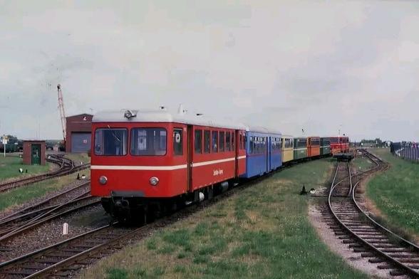 Eine kunterbunte Zusammenstellung aus Klein Triebwagen und Beiwagen stehen auf einer Abstellanlage des Inselbetriebshofs Langeooge unter freiem Himmel. Im Hintergrund sind ein kleines Wärterhäuschen und eine Werkshalle erkennbar.