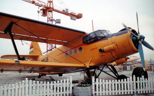 Side view of a yellow, single propellor-engined bi-plane airliner parked facing to the right, with a low white picket fence around it.
A large orange crane looms over the background, against a cloudy grey sky.