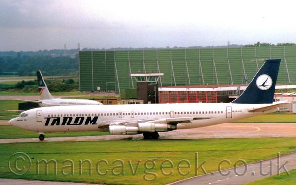 Side view of a white, 4 engined jet airliner taxiing from right to left. The plane has large blue "Tarom" titles on the lower forward fuselage, and a blue tail with a white circle containing a blue flying bird in the middle. Green grass fills the foreground, with the upper fuselage and tail of a grey jet airliner in the background on the left, and a brown brick and red metal building on the right, with a large, box-like green metal stucture behind, and trees in the distance, under grey skies.